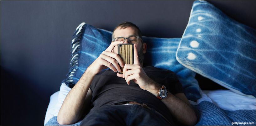 mand liggende på sengen og kigger på en smartphone
